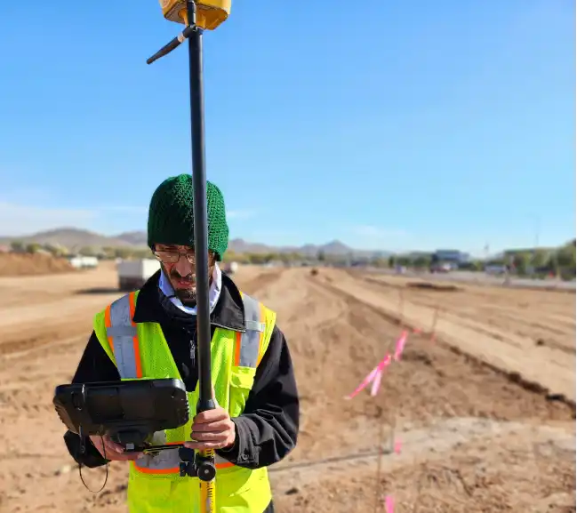 Site-engineer-operating-his-touch-screen-controller-instrument-during-roadworks (1) A construction worker in a reflective vest holds a surveying instrument on a long, cleared dirt road marked with pink flags under a clear sky